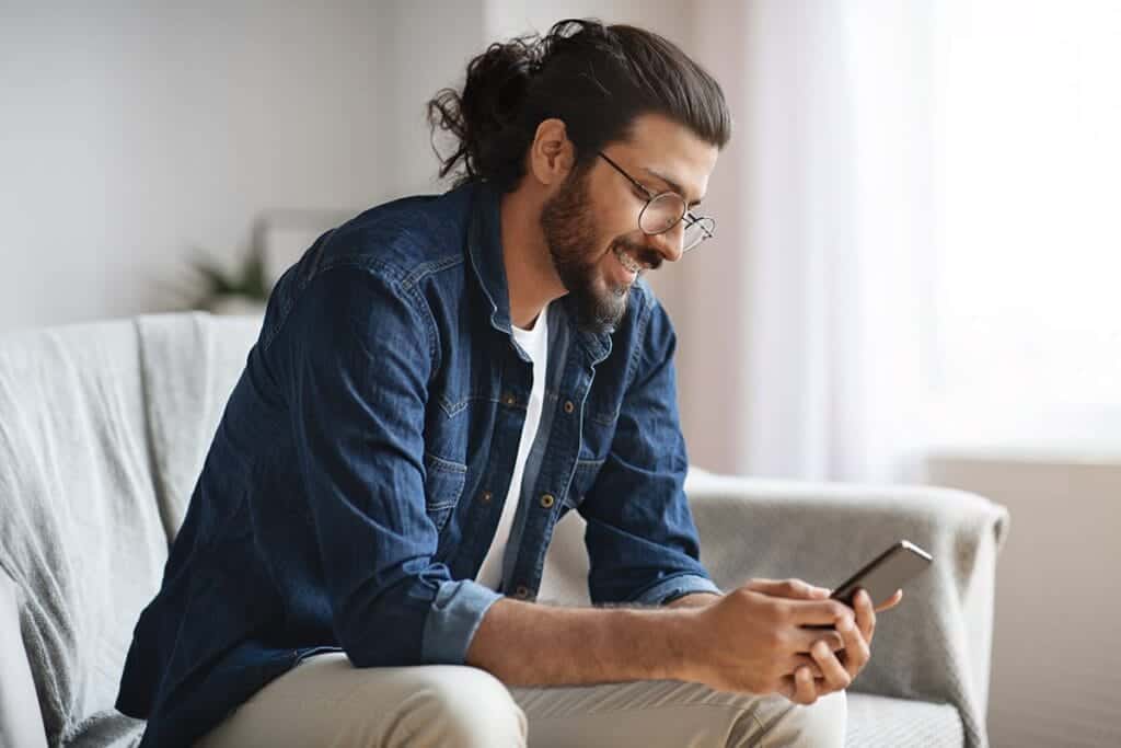 Man smiles while sitting on couch and playing on his phone while waiting to hear about alcohol treatment